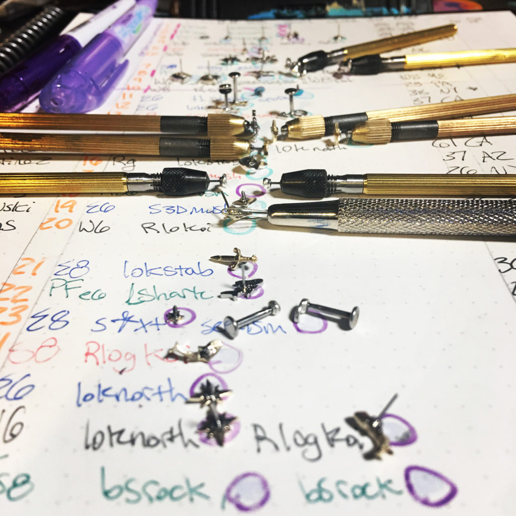 An image of a notebook with custom earring pieces set on it to match up to their orders. Many are in pin vices for polishing